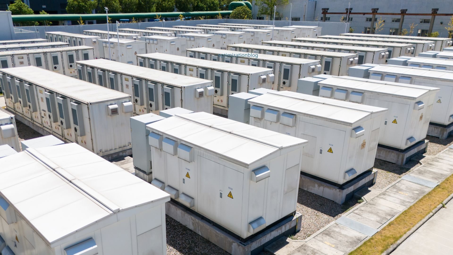 Aerial view of rows of white industrial power generators and electrical enclosures with high-voltage warning labels, arranged in a fenced outdoor equipment yard.